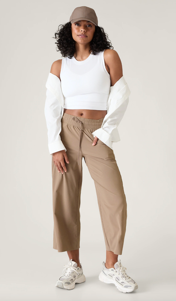 Woman in a white cold-shoulder crop top, tan trousers, and sneakers, posing for a stylish look