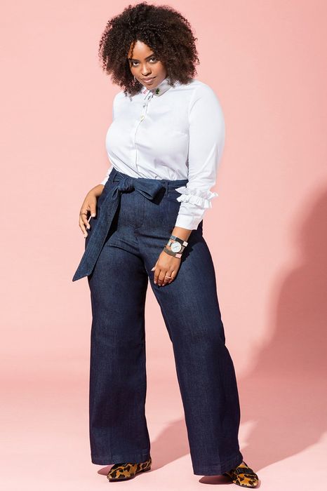 Model showcasing dark wash, wide-leg jeans with an integrated belt, standing in front of a pink backdrop.