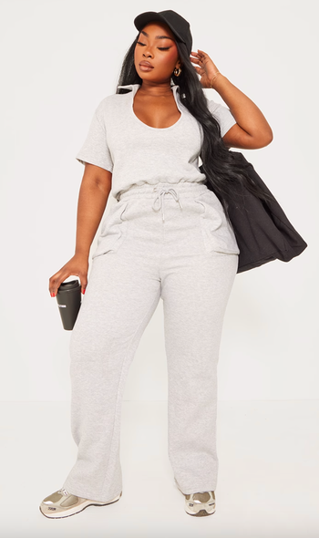 Model in casual gray loungewear, holding a black bag and a coffee cup, styled with a cap and sneakers