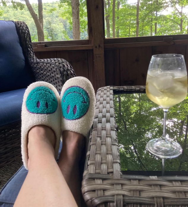 Feet adorned in snug smiley-faced slippers rest on a wicker table next to a glass of white wine, with a peaceful forest backdrop in view.