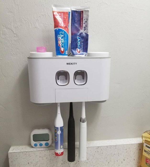 A reviewer's photo of the wall-mounted station featuring three toothbrushes hanging below and two toothpaste tubes inserted at the top