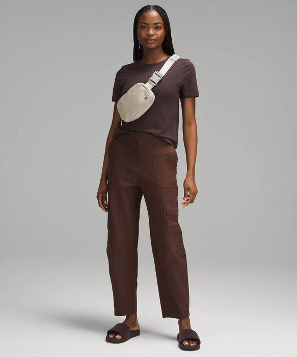 A woman confidently stands wearing a relaxed outfit featuring a dark top, matching pants, a light-colored crossbody bag, and slides.