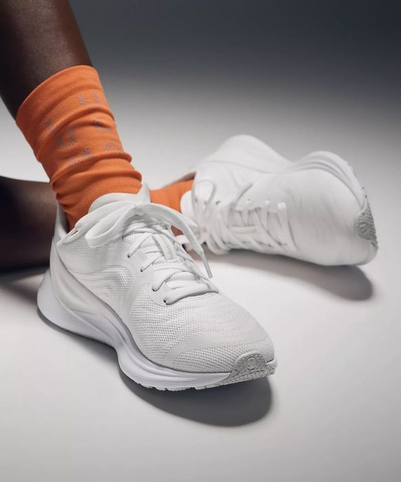 White athletic sneakers paired with orange socks featuring subtle patterns or logos, displayed on a model’s feet. Perfect for shopping or casual wear.