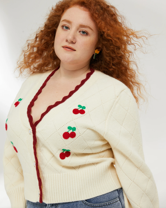 A model is wearing the cropped sweater in white with a red hem, featuring embroidered cherries on it.