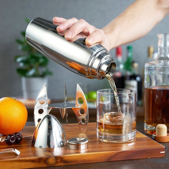 A person pouring liquid from a cocktail shaker into a glass, surrounded by various cocktail-making tools and ingredients.