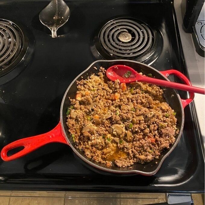 A skillet filled with ground meat and vegetables on a stove, beside a red spoon.