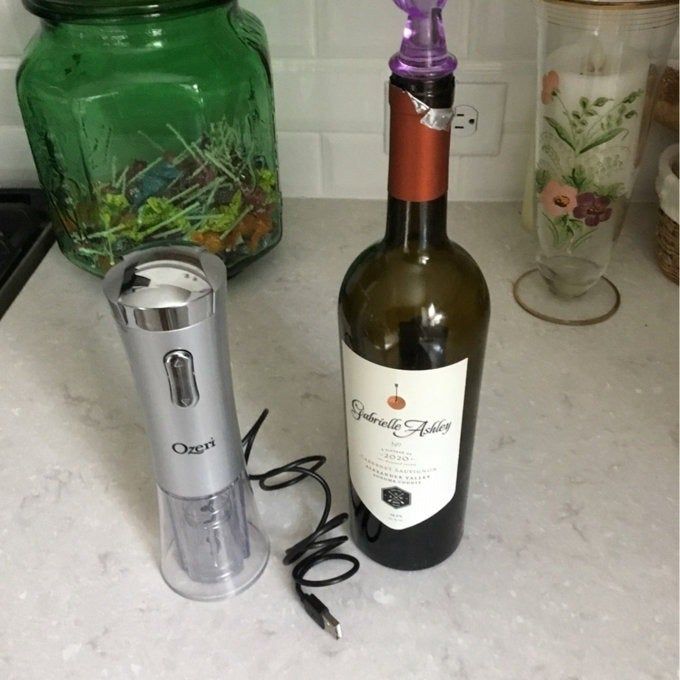 An electric wine opener beside a bottle of wine with a corkscrew twisted into the cork.