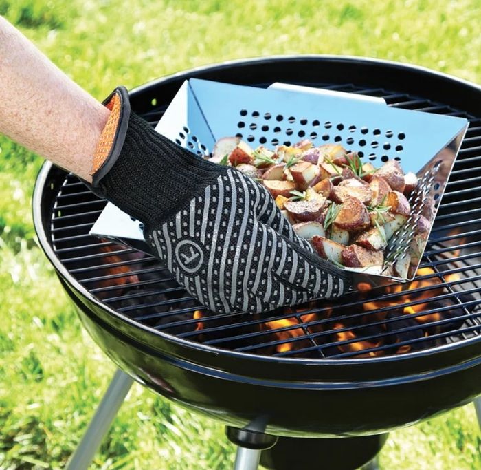 A model wearing black and gray grill gloves, holding a pan of potatoes over a hot black grill.