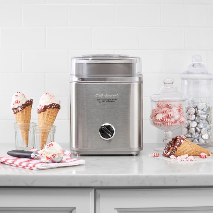 A Cuisinart ice cream maker sitting on the counter, with two cones, each topped with a scoop of ice cream, placed beside it.