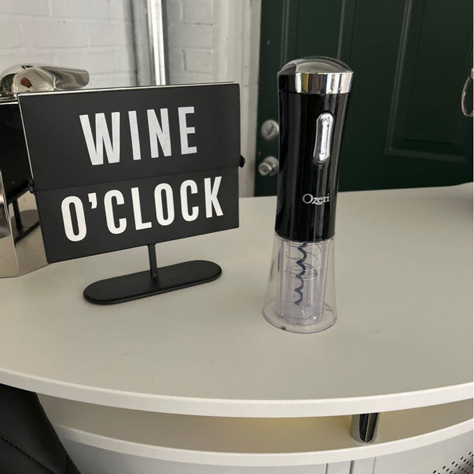 An electric wine opener placed next to a 'WINE O'CLOCK' sign on a table, making it the perfect product for wine lovers.