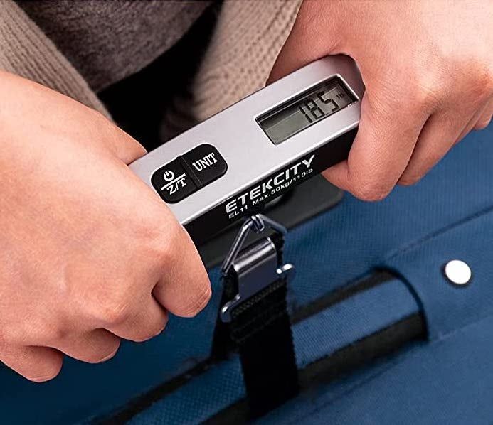 Hands holding a silver luggage scale to weigh a packed suitcase