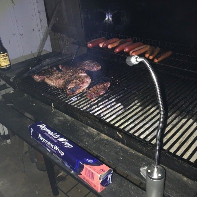 A barbecue grill with sizzling steaks and hot dogs, a box of Reynolds Wrap, and a grill light attached to ensure perfect visibility while cooking.