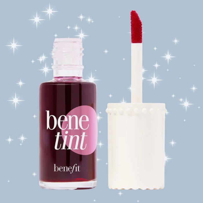 A bottle of Benefit Cosmetics Benetint, a versatile lip and cheek stain, shown with its applicator removed and placed beside it, surrounded by sparkling effects.