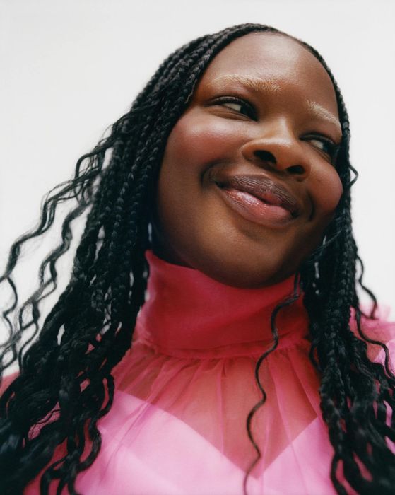 Close-up of a person smiling in a sheer pink blouse with a ruffled collar, sporting long braids.