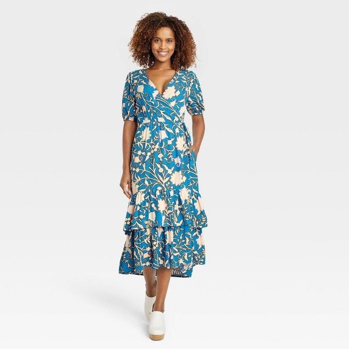 A model is showcasing a blue wrap dress adorned with white and green floral patterns, paired with matching green shoes.