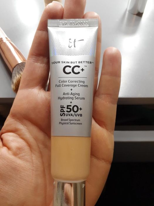 A close-up of a hand holding a tube of IT Cosmetics CC+ Color Correcting Full Coverage Cream with SPF 50+, mentioning anti-aging hydrating serum and physical sunscreen on the packaging.
