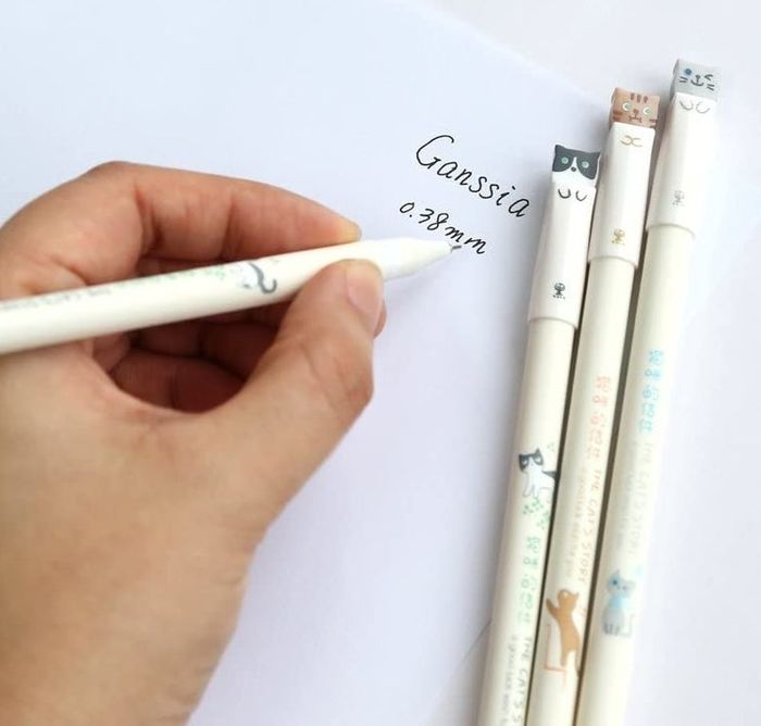 A model is shown writing on paper with black ink, accompanied by white pens featuring cat-shaped tips.