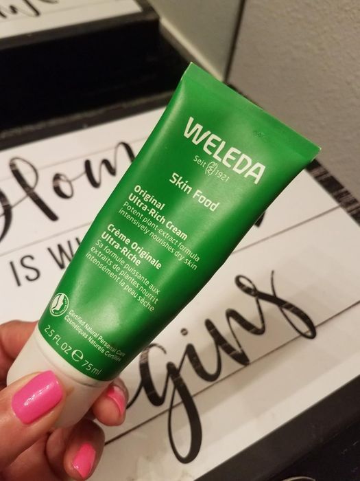 A close-up shot of a hand holding a tube of Weleda Skin Food Ultra-Rich Cream, with part of a sign in the background partially visible.