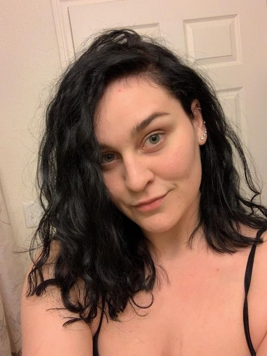 A woman with curly, shoulder-length hair and minimal makeup poses for a selfie in a bathroom, wearing a black strap top.