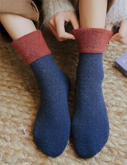 A person lounging in cozy knit socks, one pair featuring a red cuff, relaxing on a woven rug.