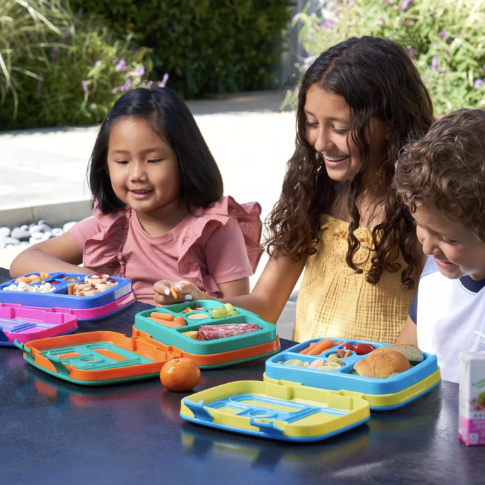Children happily eating from colorful bento lunch boxes; one girl wears a pink dress, another has a yellow top, and a boy is dressed in a blue and white striped shirt