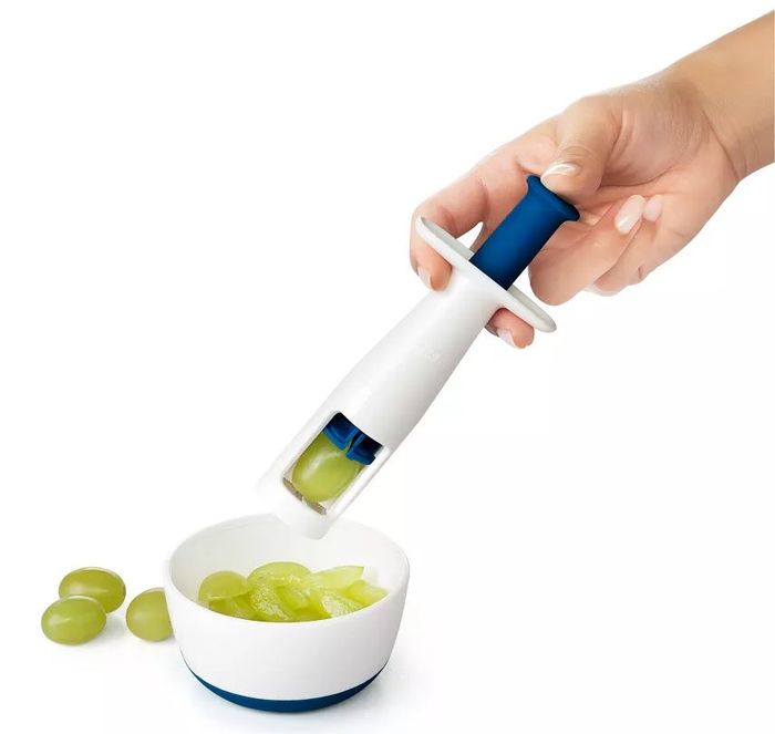A hand is seen using an OXO Good Grips grape cutter to slice green grapes into a bowl