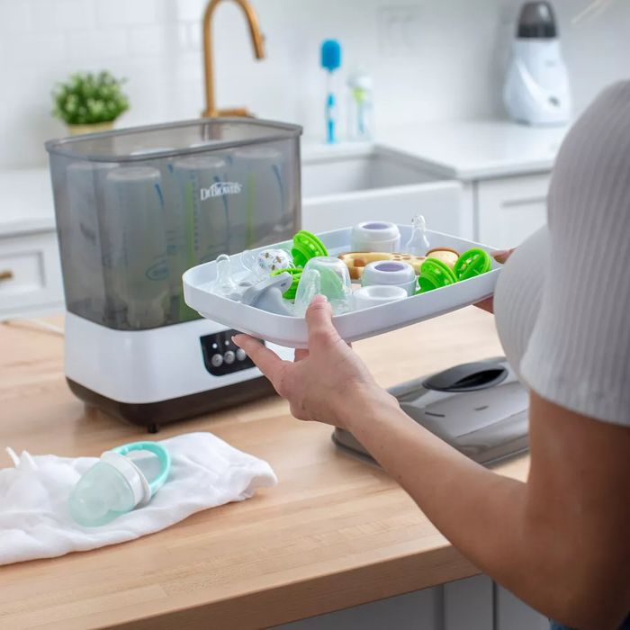 A person is using a bottle sterilizer, with various baby items spread out across the kitchen counter.