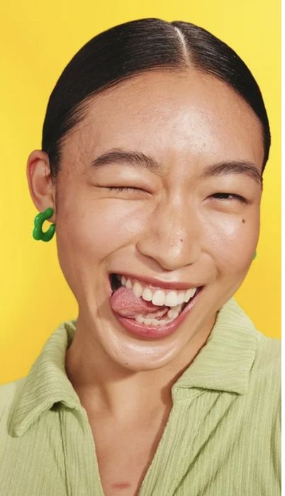 A person playfully winks and smiles while wearing green earrings and a light-textured top, radiating cheerful energy.