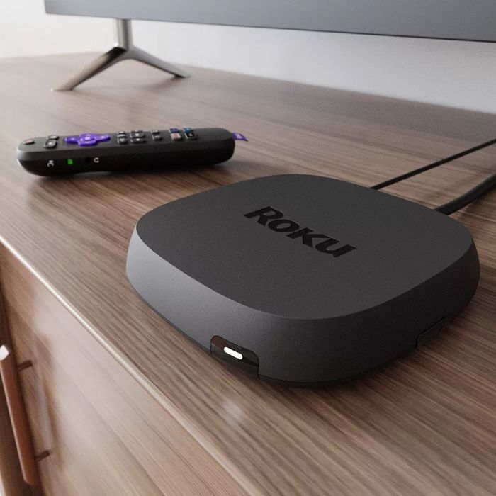 A Roku streaming device rests on a wooden entertainment unit, accompanied by a remote control, with a partially visible TV screen in the background