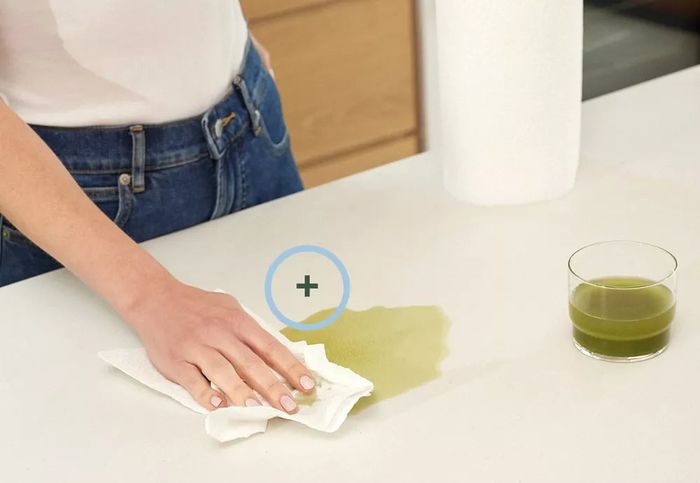 A person cleaning a green liquid spill on a countertop using a paper towel, with the text: 