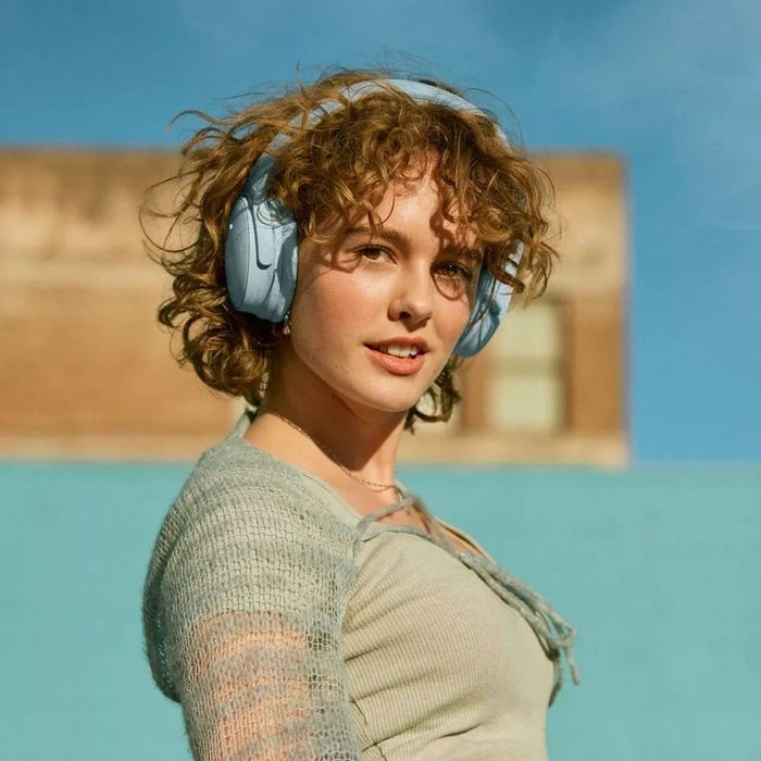 A person with curly hair, wearing large headphones and a cozy, light knitted sweater, smiles at the camera while enjoying the outdoors. This moment is featured in an article focused on headphone shopping.