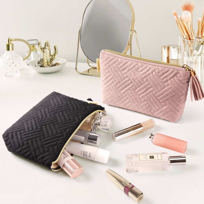 Two quilted makeup bags, one in black and the other in pink, rest on a vanity surrounded by various beauty products such as lipsticks and perfume.
