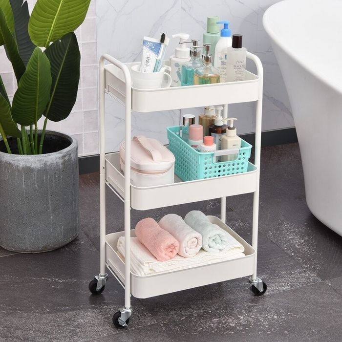 A bathroom storage cart on wheels, featuring three shelves neatly organized with toiletries, towels, and hygiene products. The cart is positioned next to a potted plant and bathtub.