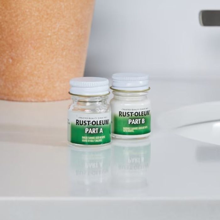 Two small containers labeled Rust-Oleum Part A and Part B are positioned on a counter beside a sink and a plant pot.