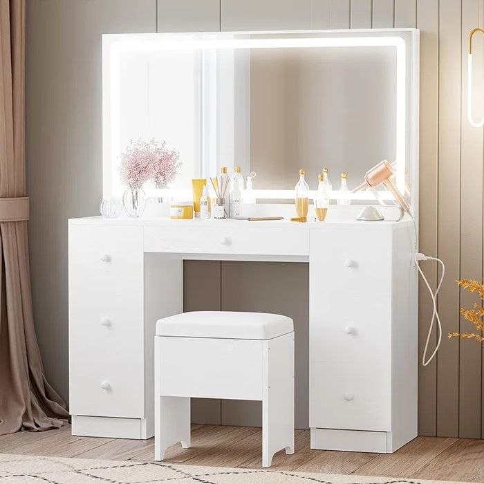 A white vanity table features a backlit mirror, a selection of cosmetics, and a bouquet of flowers placed on top. A white stool is positioned in front of the table.