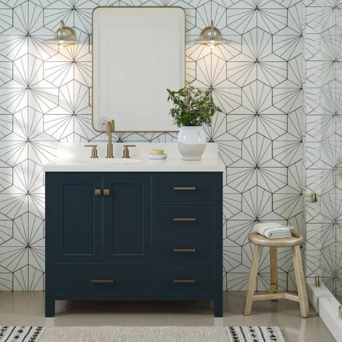 A contemporary bathroom featuring geometric wallpaper, a rectangular mirror, twin light fixtures, a dark vanity with brass accents, and a small potted plant on the counter.