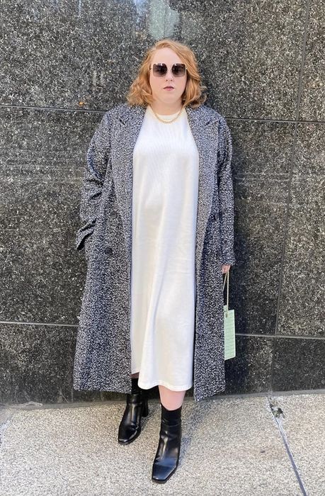 Gillian Jacobs poses in front of a marbled wall, wearing a long textured coat over a white dress, paired with black ankle boots, sunglasses, and a gold necklace