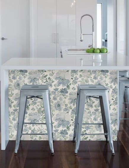 A contemporary kitchen featuring a white central island, adorned with floral-patterned wallpaper, complemented by two sleek gray metal stools and a modern faucet. A bowl of apples adds a touch of freshness on the countertop.