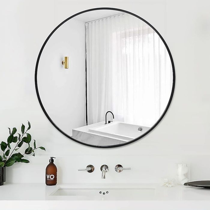 A contemporary bathroom featuring a round mirror reflecting a bathtub and white curtains. The counter is adorned with a plant and a VO soap bottle.