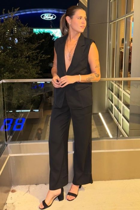 A reviewer standing on a balcony at night, wearing a sleeveless black suit paired with heels, gazing into the distance