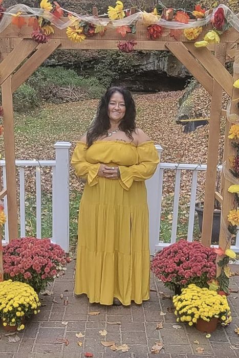 A reviewer stands beneath a flower-draped wooden arch, dressed in a flowing off-shoulder dress, surrounded by potted chrysanthemums on a brick path.