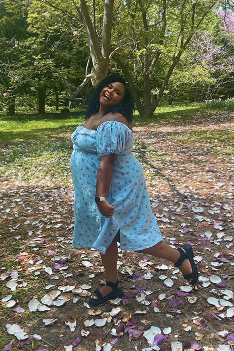 A reviewer smiles while standing in a park surrounded by fallen petals, wearing a floral dress with one hand resting on her hip