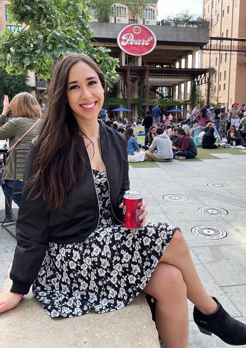 The reviewer is seated outside, clutching a can, dressed in a floral-patterned dress paired with a black jacket.