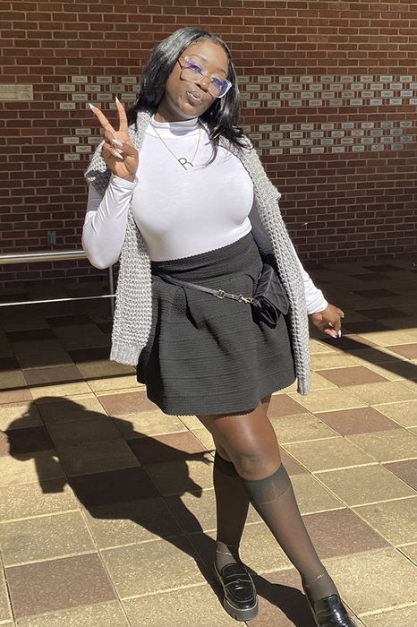 A person joyfully poses with a peace sign, wearing a white turtleneck, gray knitted vest, black skirt, knee-high socks, and loafers.