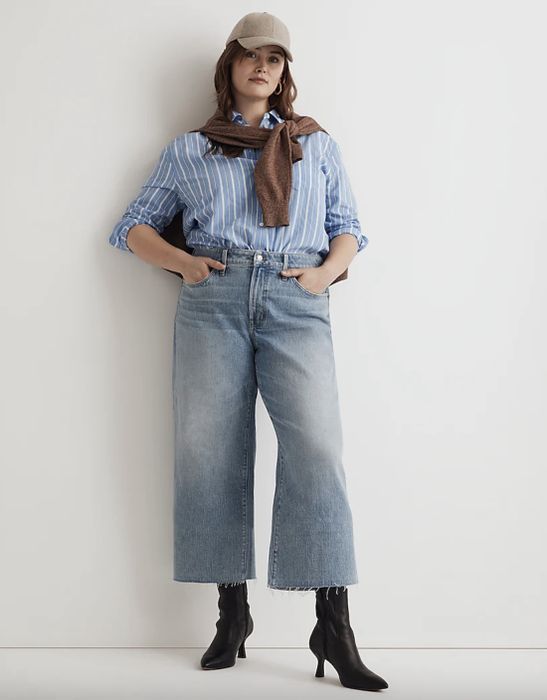 A woman in a striped shirt, jeans, and boots, accessorized with a scarf and hat, posing with hands in her pockets.