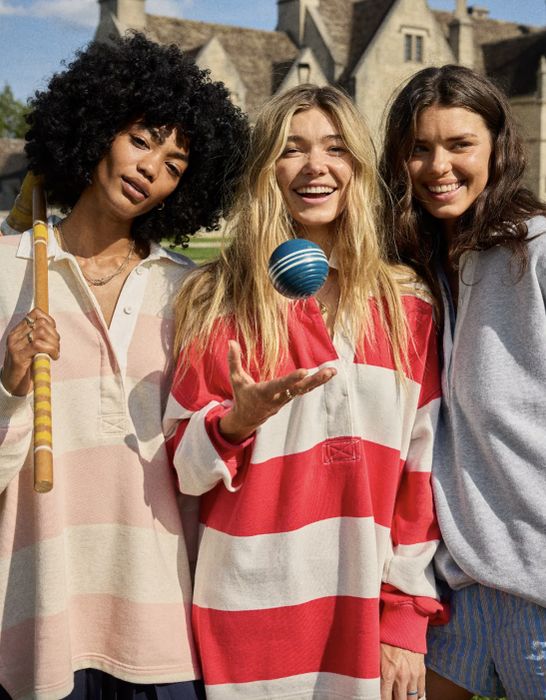 Three women, all smiling and standing together outdoors, one holding a striped croquet mallet. They are dressed in casual, oversized striped shirts.