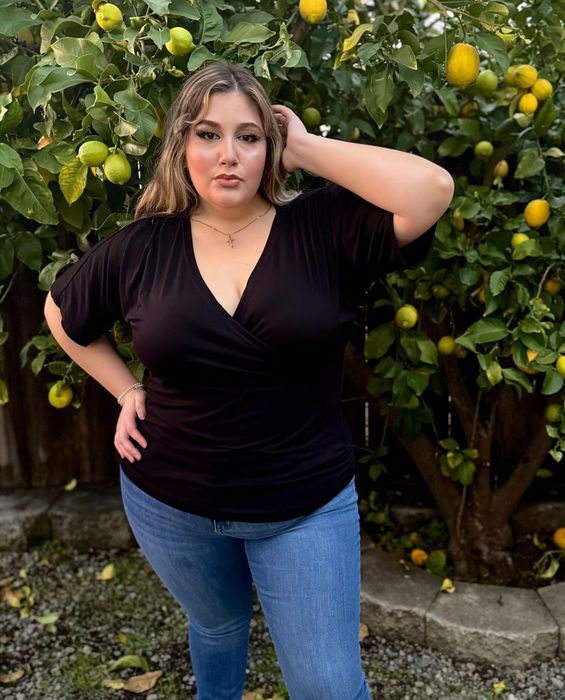 A reviewer poses outdoors in a black top and jeans, standing in front of a lemon tree with their hands placed on their head and hip