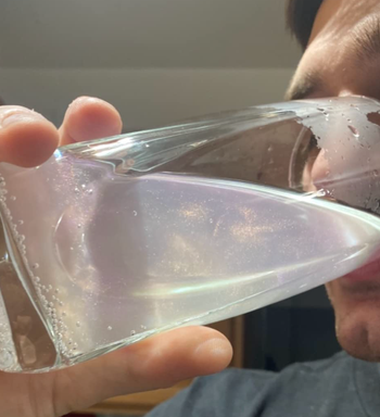 A person sipping from a clear glass filled with shimmering liquid.