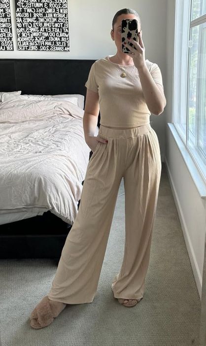 A person in a mirror selfie wearing a casual loungewear set consisting of a T-shirt and wide-legged pants, one hand in their pocket, one foot wearing a slipper. The background features an unmade bed.