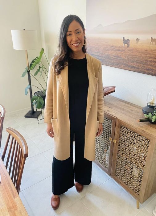 A woman smiles, standing in a room with chic decor, wearing a long beige coat over black attire and brown shoes. Small plants and a horse picture decorate the background.